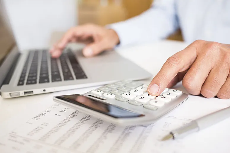 Person with a laptop and calculator at a desk, sheets of paper with figures on next to them