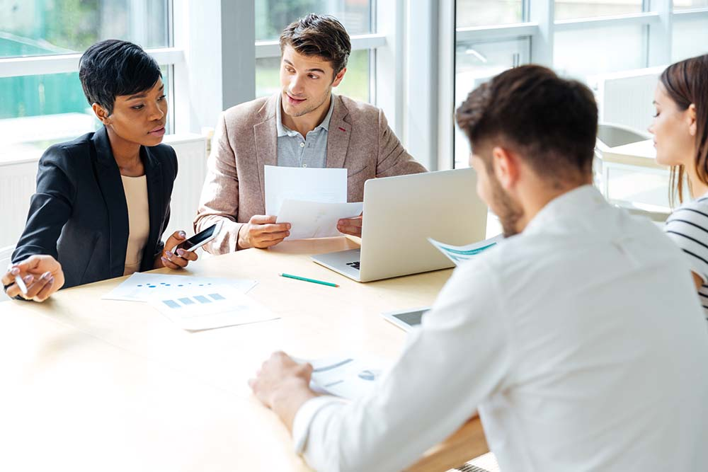 Two ladies and two men sat around a table in an office scenario with laptops and papers