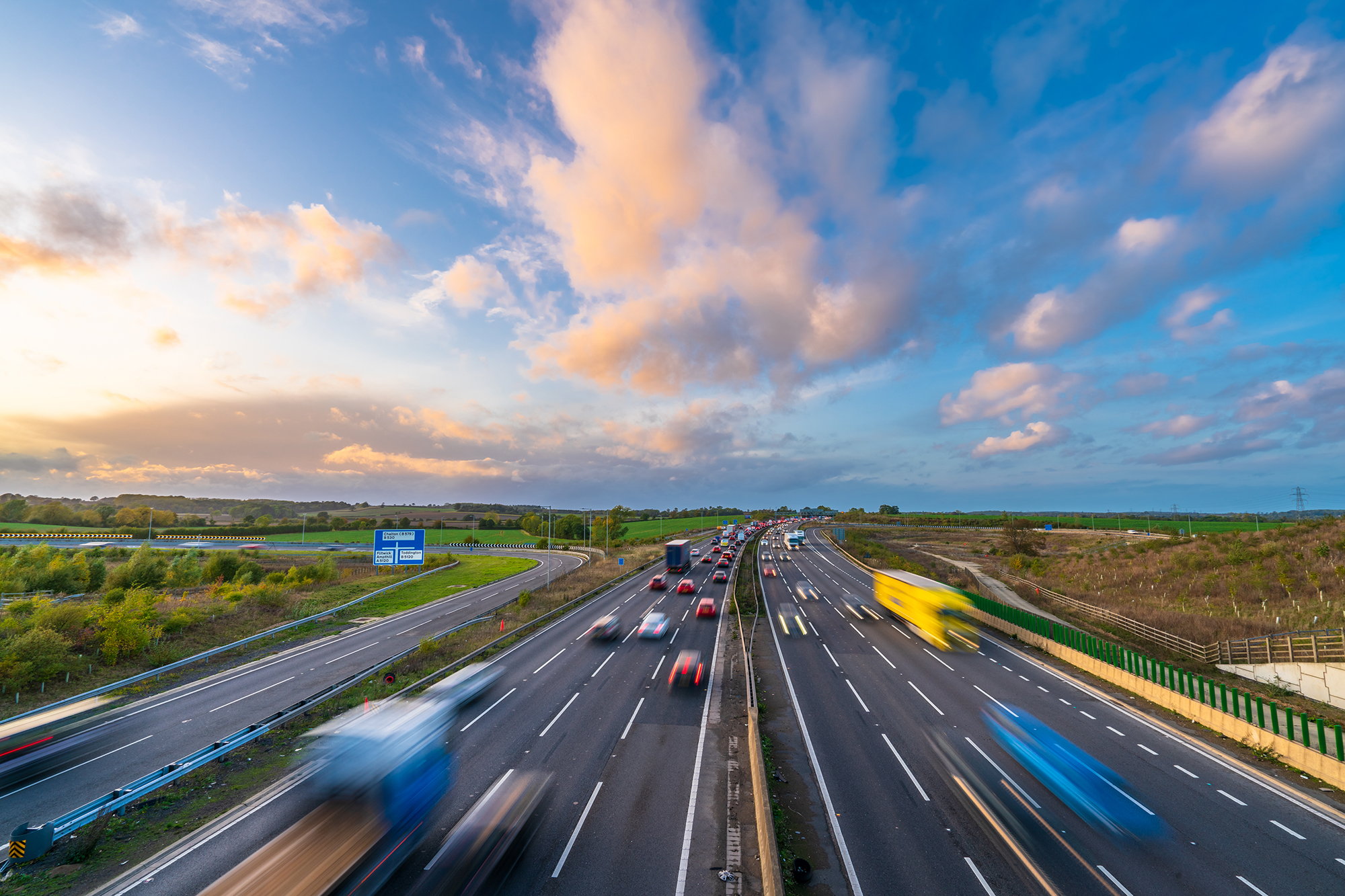 A motorway shown with multiple vehicles going in both directions, from a high up view point, blue sky and clouds at the top of image